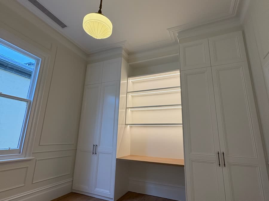 A period room with an ornate ceiling rose, a globe pendant and strip-lit joinery shelves
