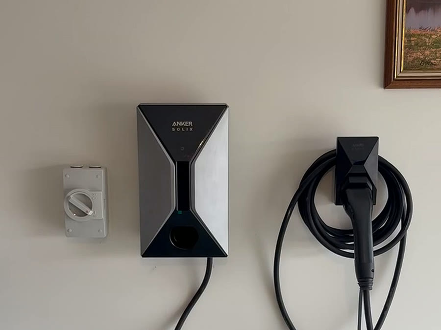 A wall-mounted home EV charger with its cable coiled on the holster and an isolator switch beside it