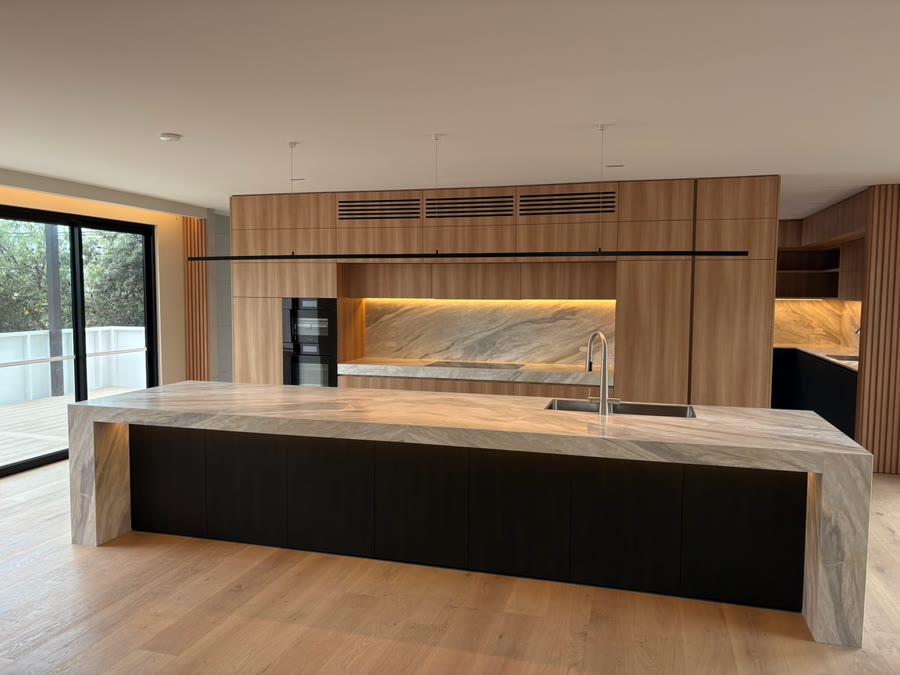An open-plan kitchen with a stone island bench, concealed strip lighting under the joinery and a linear pendant running the length of the room
